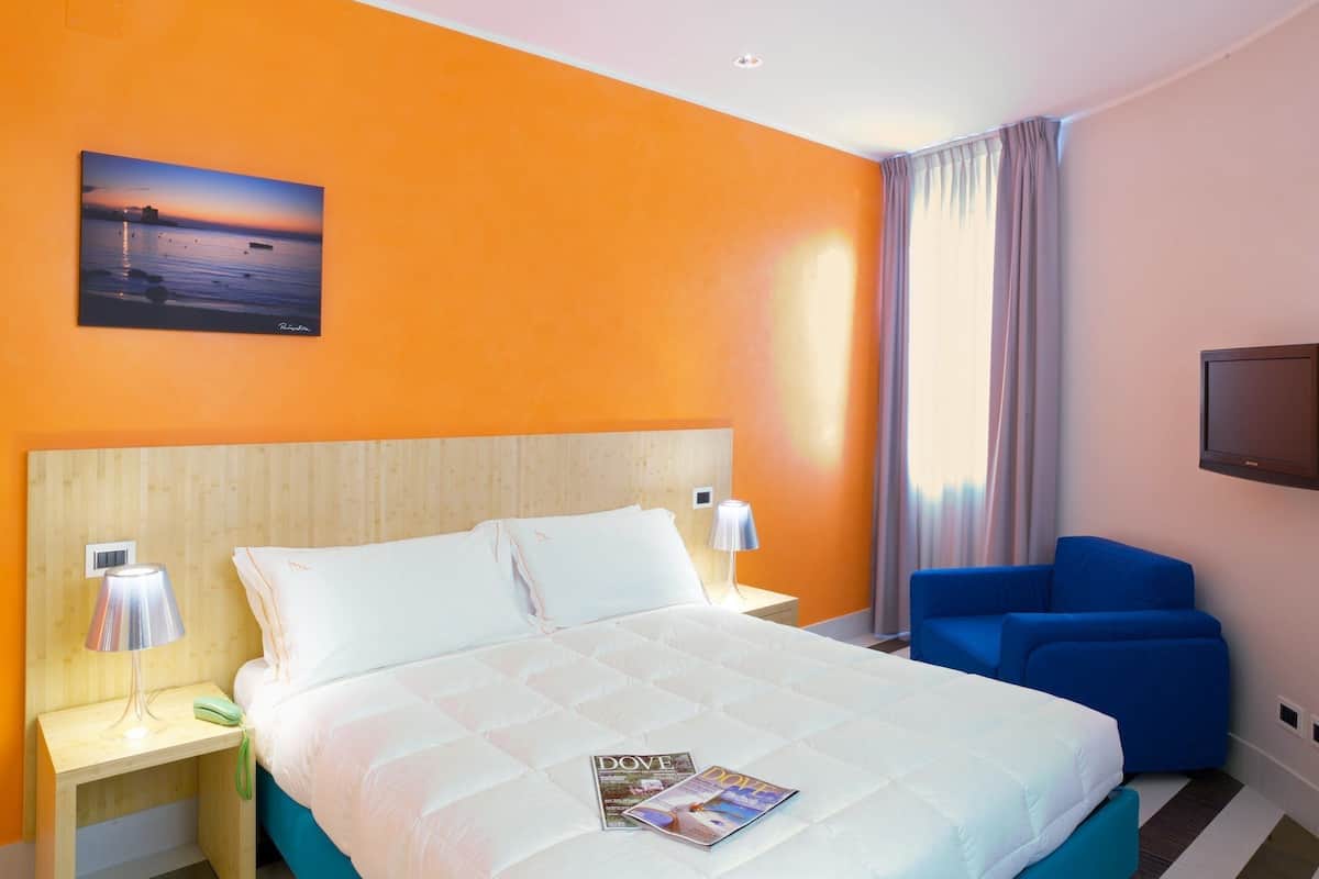 Habitación de hotel con una cama grande, una mesita de noche con lámpara, una silla azul, un televisor y un cuadro de un atardecer en la pared.
