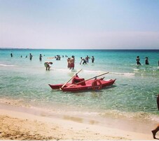 A beach with clear turquoise water, a red boat, and people swimming and standing in the water.