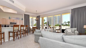 A modern living room with a grey sofa, wooden chairs, a dining table, and a kitchen area with cabinets and a microwave. - Large lobby-level end condo w/patio access to beach & pool! (Destin)