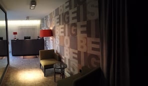 Habitación de hotel con diseño moderno, que presenta una pared grande con texto repetido, un sofá, una mesita redonda y un escritorio con lámpara.