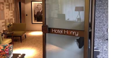 Un hall d’accueil d’hôtel avec une porte en verre portant l’inscription « Hôtel Henry », un fauteuil vert, une petite table sur laquelle est posé un vase de fleurs et un tableau encadré accroché au mur.