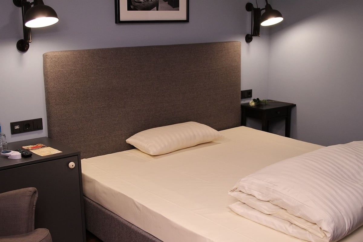 Cama bem arrumada com cabeceira, mesa de cabeceira com abajur e criado-mudo com luminária.
