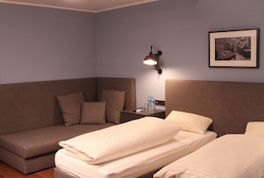 Habitación de hotel con sofá, dos camas, una mesita de noche con lámpara y un cuadro en la pared.