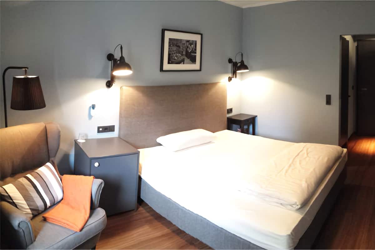 Quarto de hotel moderno com cama, abajures, mesa de cabeceira, sofá e um quadro na parede.