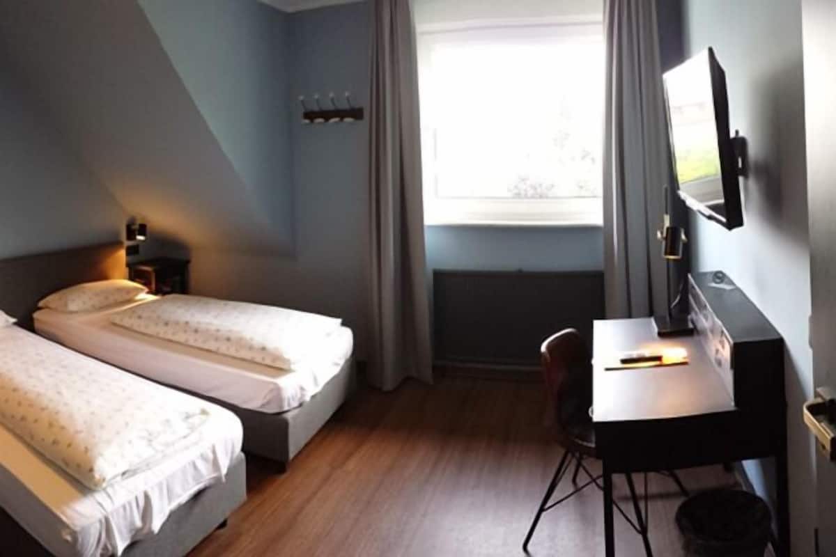 Quarto de hotel com duas camas de solteiro, uma escrivaninha, uma cadeira, uma luminária, um quadro na parede e uma janela com cortinas.