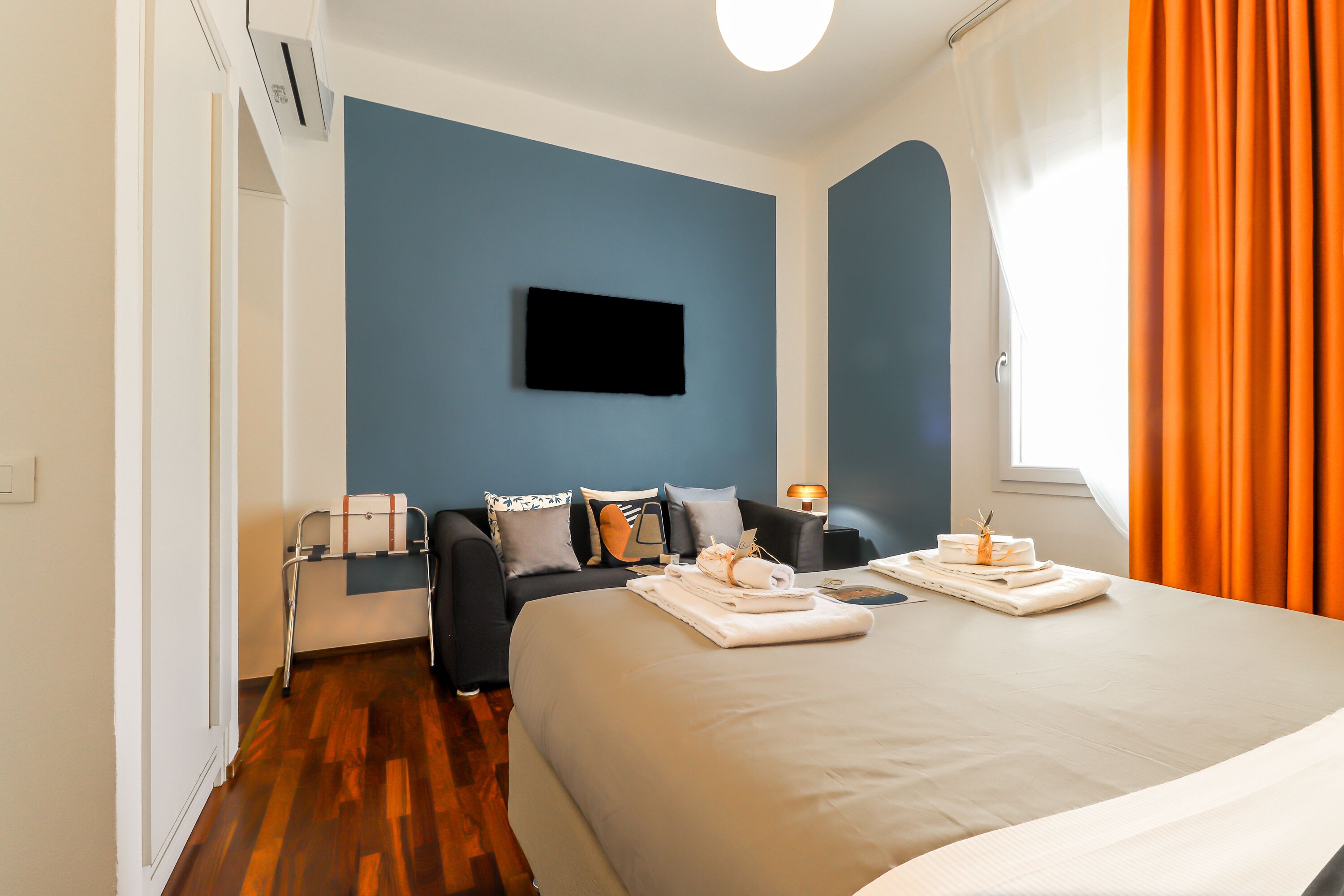 A modern hotel room with a large bed, a flat-screen TV mounted on the wall, a bedside table with a lamp, and a window with orange curtains.