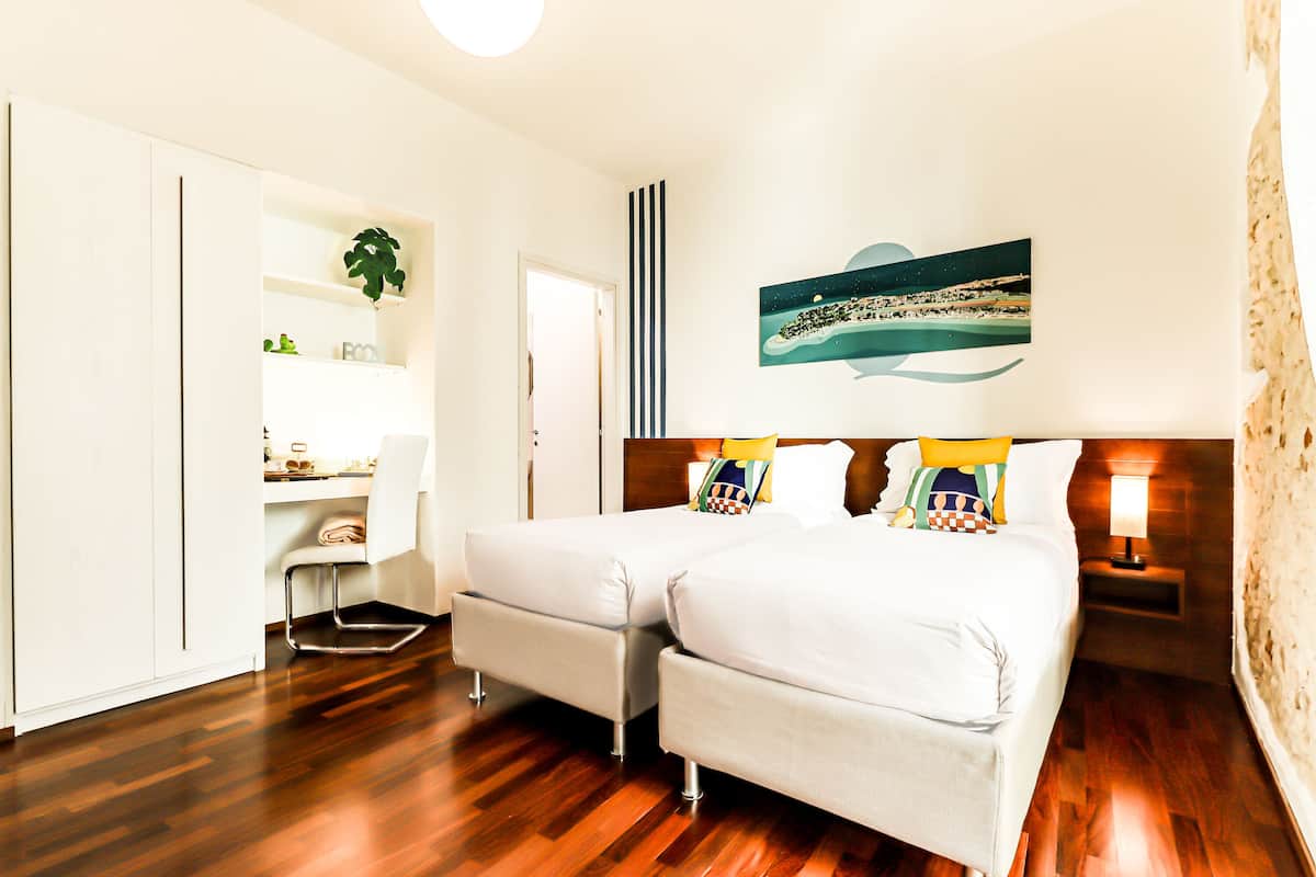 A hotel room with a wooden floor, a bed with white bedding, a desk with a chair, a plant, and a wall-mounted artwork.