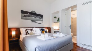 A neatly made bed with a headboard, a bedside table with a lamp, and a wall-mounted artwork depicting a night scene. - Qaral Bed and Breakfast (Cagliari)