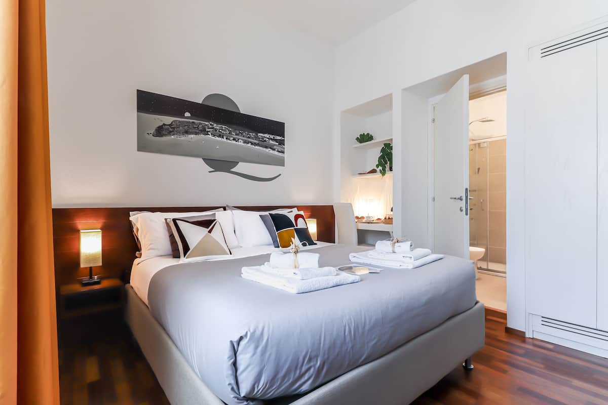 A neatly made bed with a headboard, a bedside table with a lamp, and a wall-mounted artwork depicting a night scene.