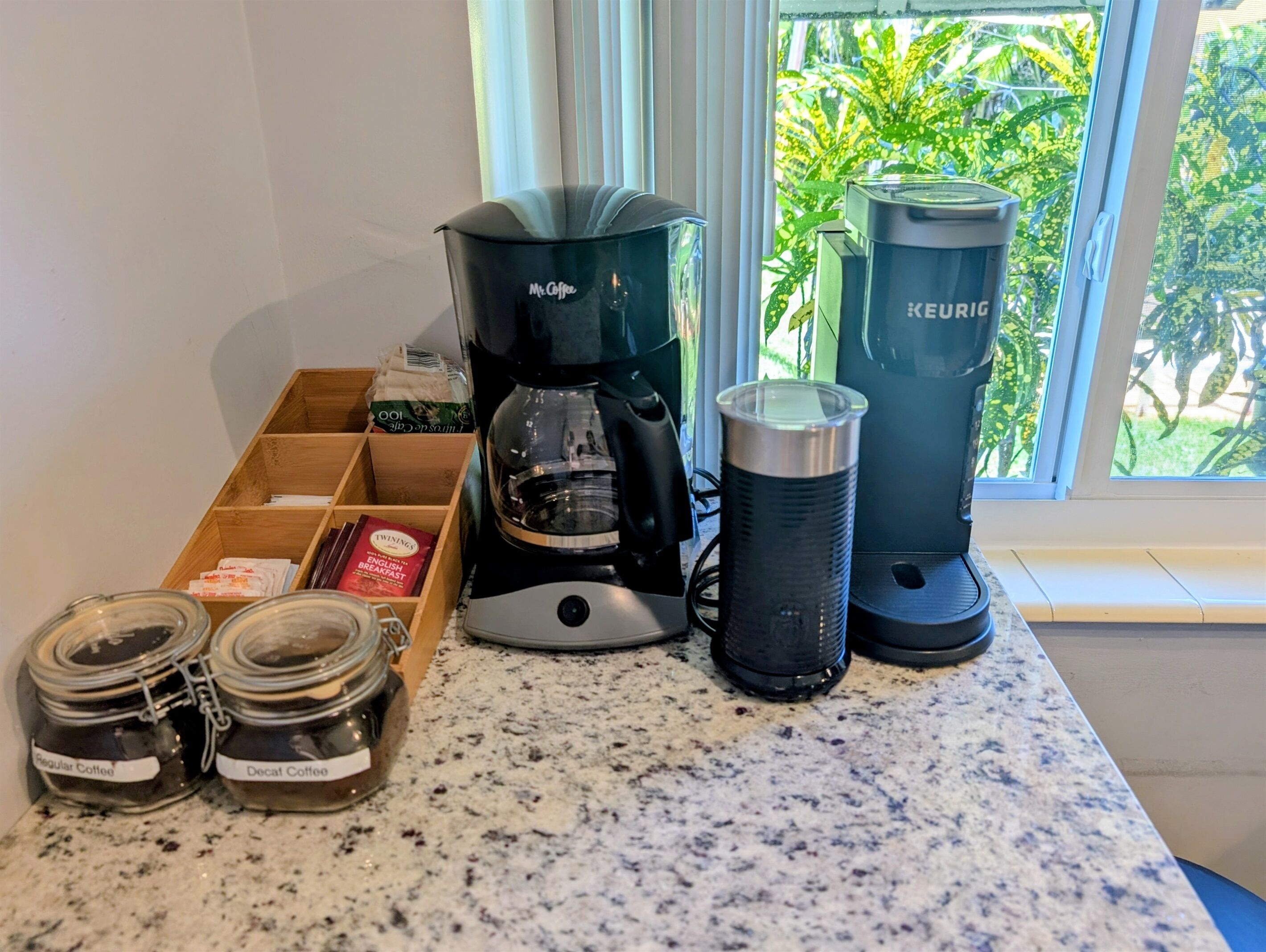 A kitchen counter with a Keurig coffee maker, a coffee grinder, and two Keurig coffee makers. There are also two jars labeled "Regular Coffee" and "Decaf Coffee."