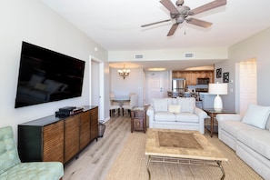 A living room with a flat-screen TV, a ceiling fan, a dining area with a table and chairs, a kitchen with wooden cabinets, and a light-colored sofa.