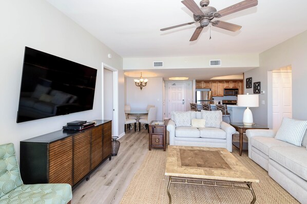 A living room with a flat-screen TV, a ceiling fan, a dining area with a table and chairs, a kitchen with wooden cabinets, and a light-colored sofa. - 2BR/2BA Condo Steps from the Beach in Destin (Destin)