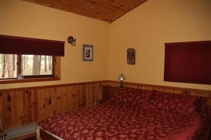 Une chambre avec un plafond en bois, un grand lit recouvert d’une courtepointe à motifs rouges, une fenêtre donnant sur les arbres et une lampe fixée au mur.