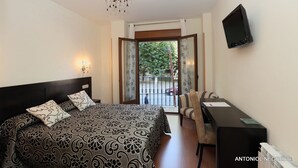 A bedroom with a bed, bedside lamps, a desk, a chair, a TV, and a balcony with a view of the street. - Hotel HC Sigüenza (Siguenza)