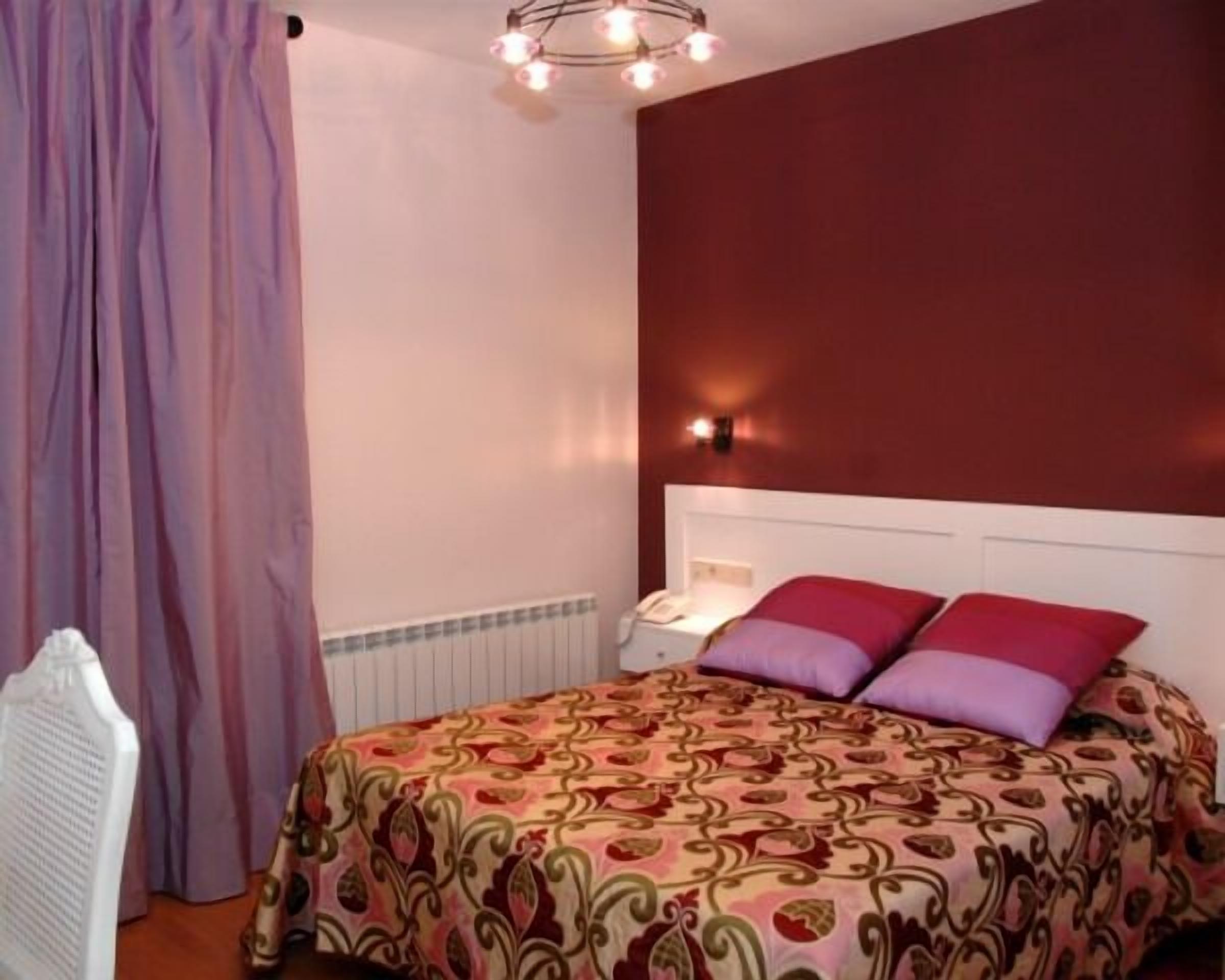 A bedroom with a patterned bedspread, a white headboard, a bedside table with a lamp, and a wall-mounted light fixture.