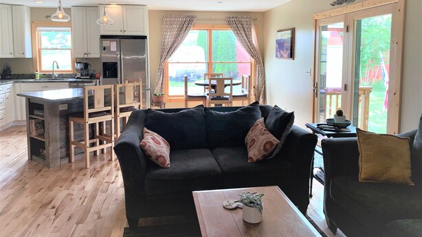 Uma sala de estar aconchegante com um sofá escuro, mesa de centro de madeira e uma área de jantar com mesa e cadeiras. Uma cozinha com armários brancos e uma geladeira é visível ao fundo.