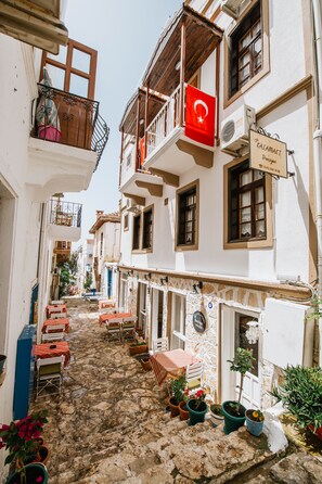 A narrow street with buildings on either side, a flag, and outdoor seating. - Old Kalamaki Pansiyon (Kas)