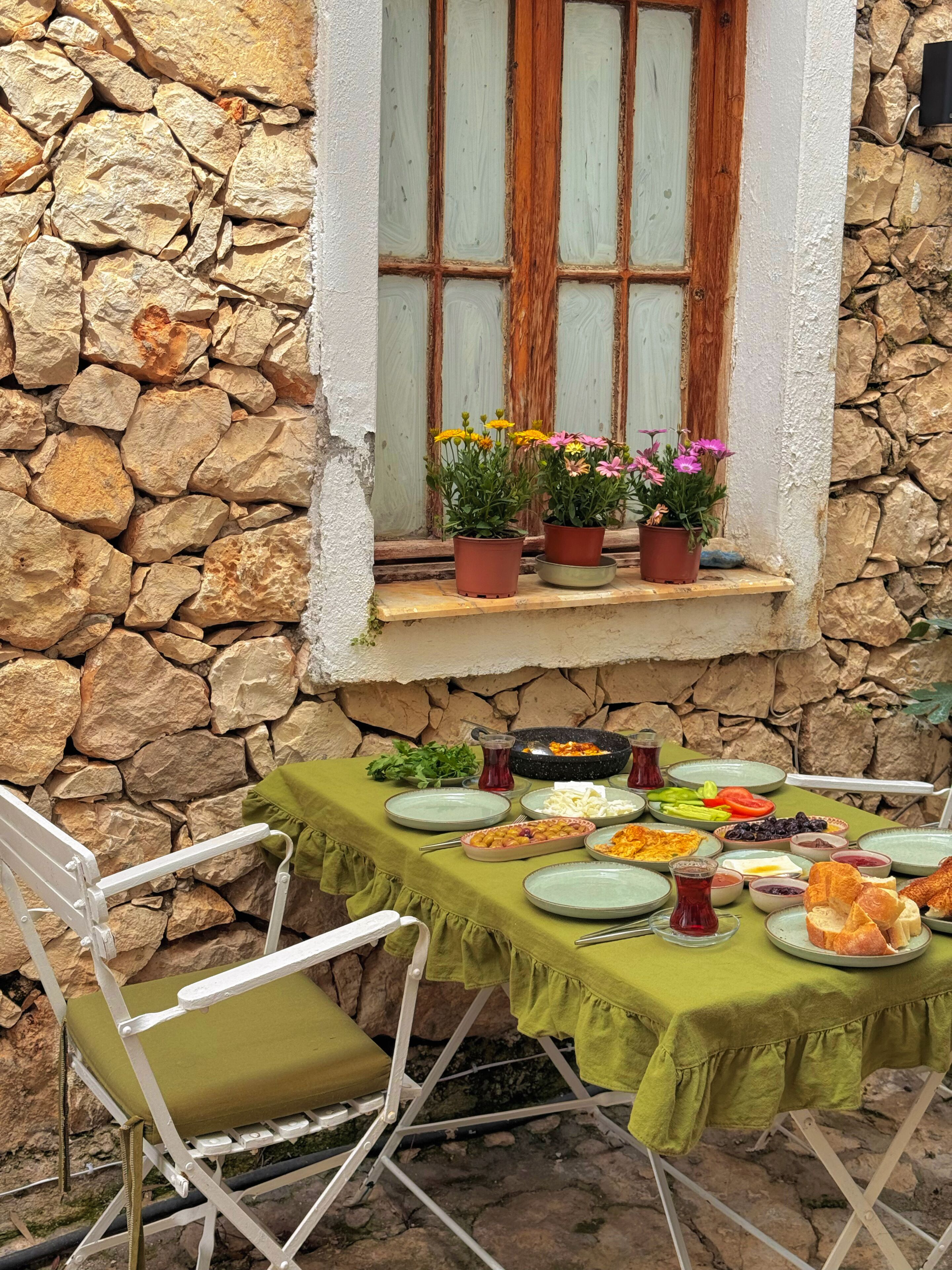 Un mur de pierres, une fenêtre avec des volets en bois, une table recouverte d’une nappe verte et des plantes en pot sur le rebord de la fenêtre.