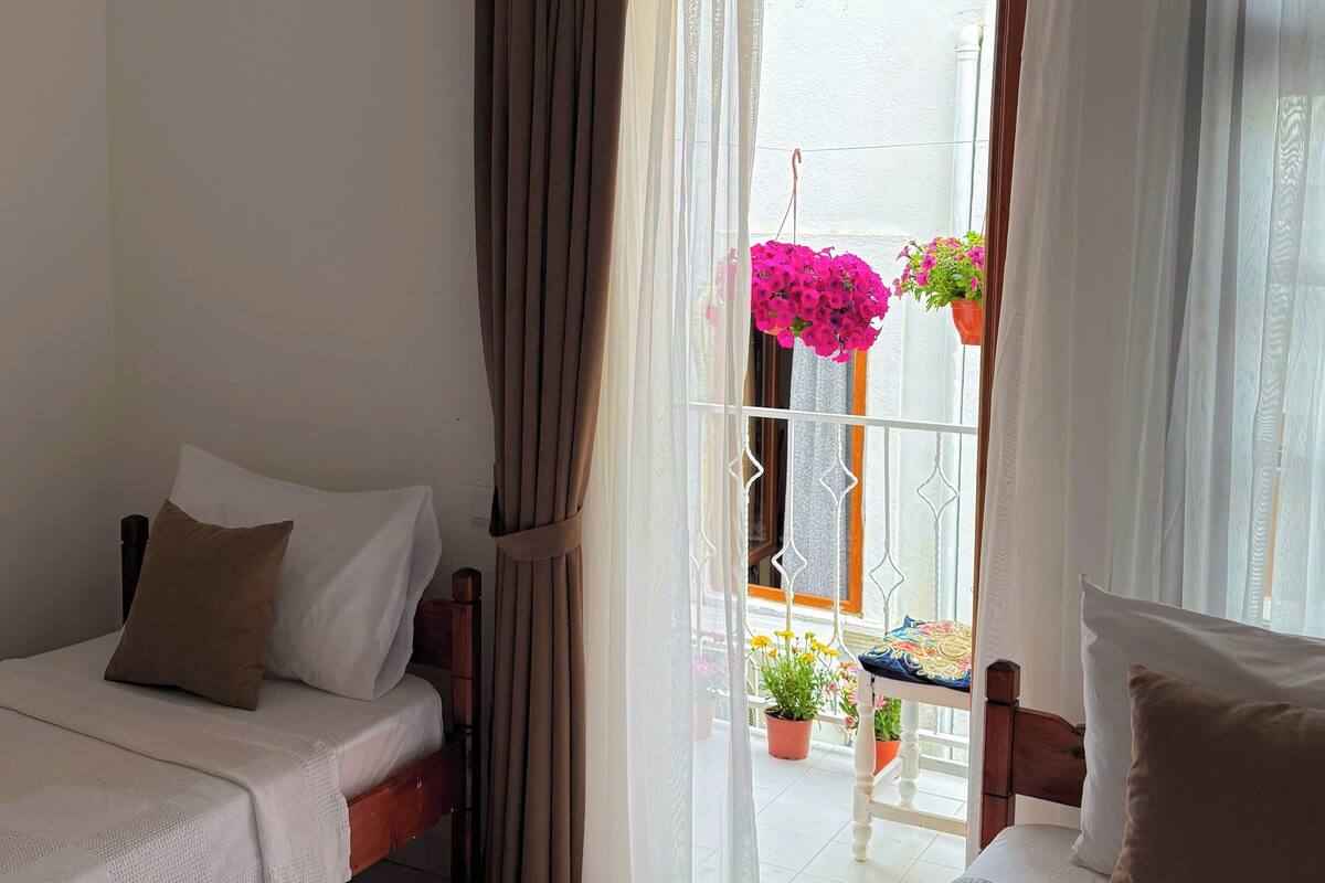 Ein Schlafzimmer mit einem Bett, einem Sessel, einem Fenster mit Vorhängen und Topfpflanzen.