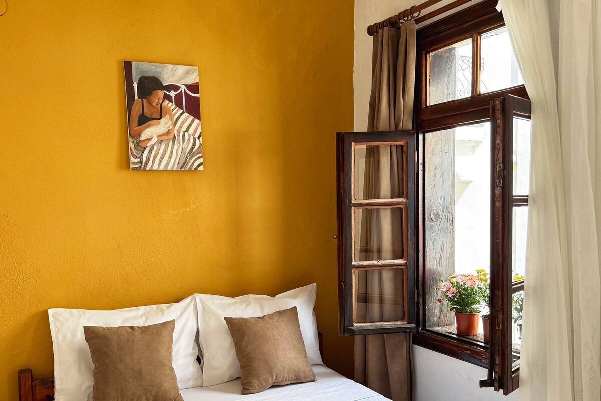 Ein Schlafzimmer mit Bett, Kissen, einem Gemälde, einem offenen Fenster mit Vorhängen und einem Blumentopf auf dem Fensterbrett.