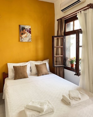 Un dormitorio con cama, almohadas, un cuadro, una ventana abierta con cortinas y una planta en el alféizar.