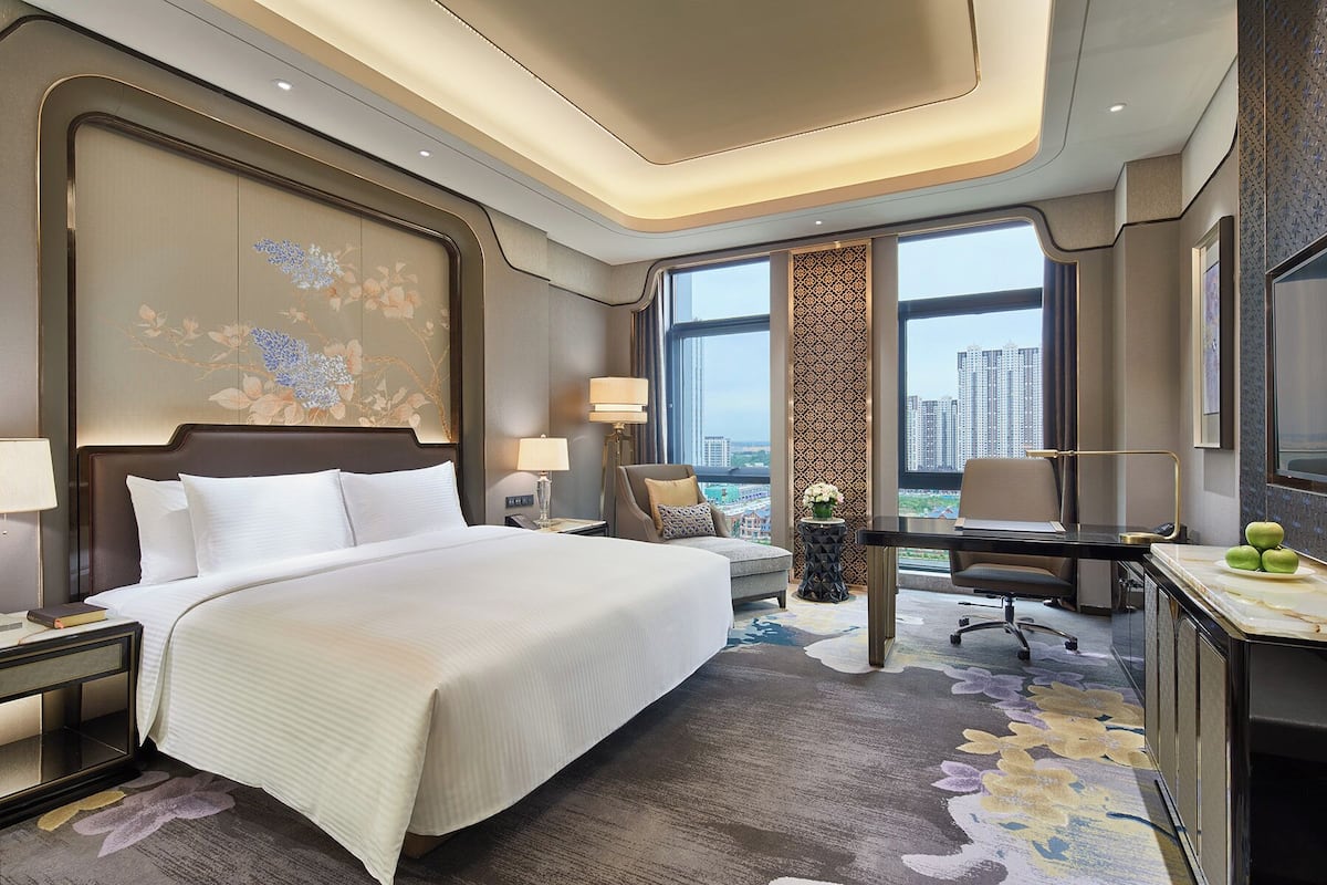 A hotel room with a large bed, a desk, a chair, and a view of the city through large windows.