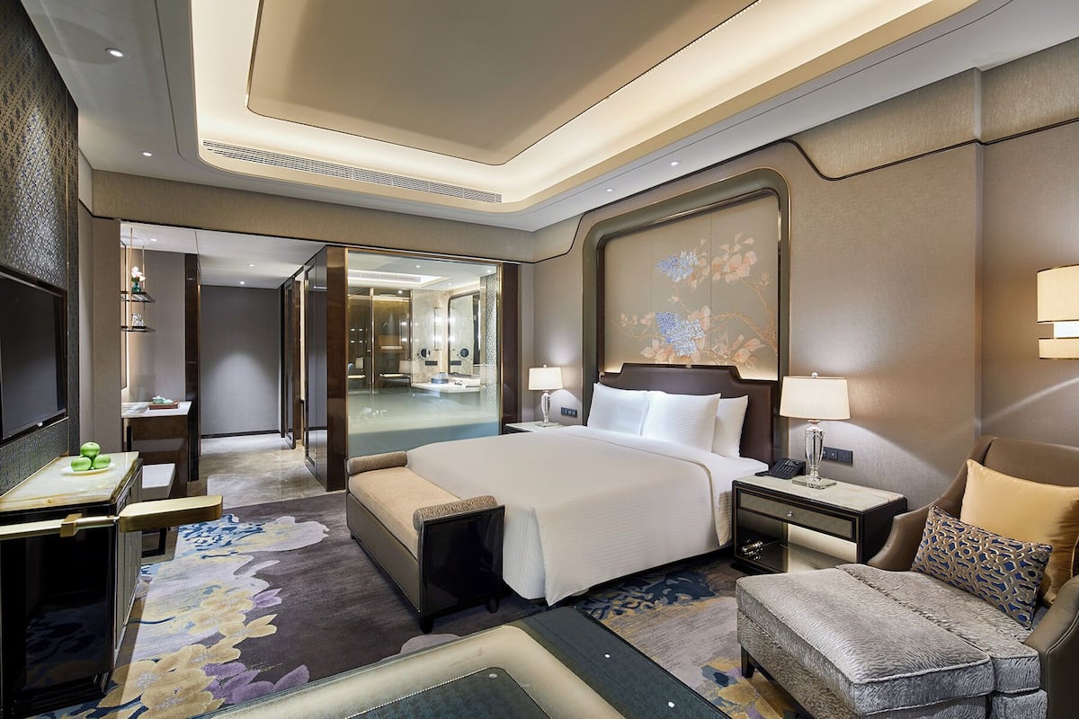 A modern hotel room with a large bed, bedside tables, a seating area, and a bathroom visible through an open door.