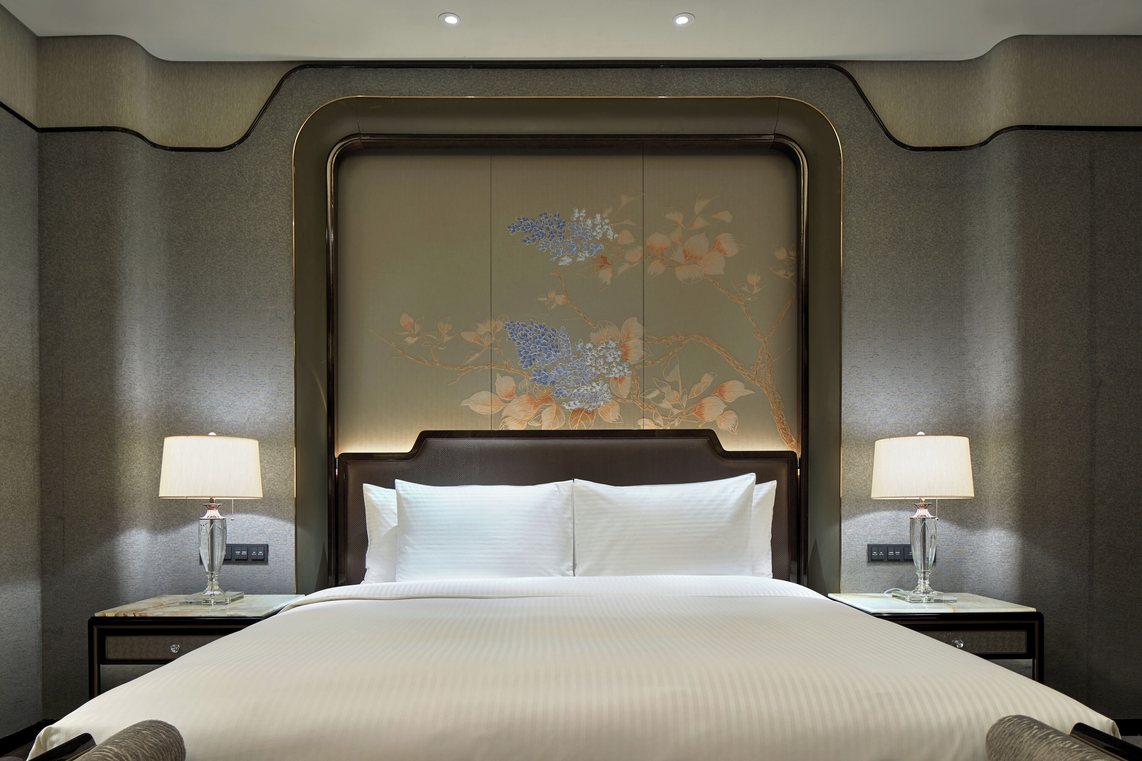 Una cama grande con sábanas blancas, flanqueada por dos mesitas de noche con lámparas, en una habitación con un cabecero decorativo y un mural floral.