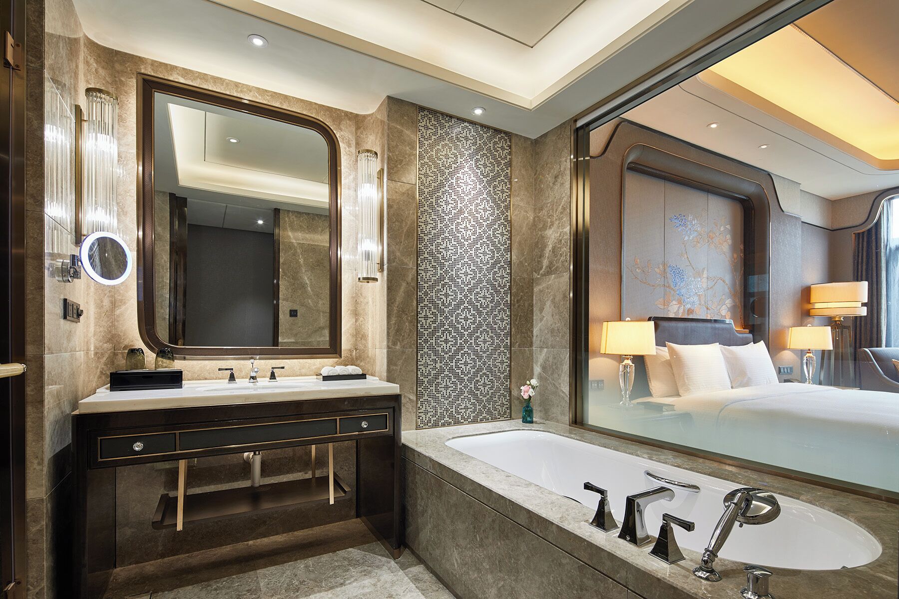 A modern bathroom with a large mirror, dual sinks, a bathtub, and a large window offering a view of a bedroom.