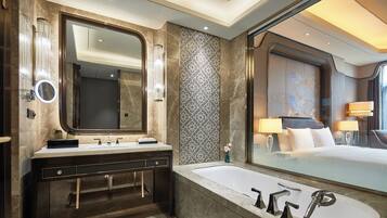 A modern bathroom with a large mirror, dual sinks, a bathtub, and a large window offering a view of a bedroom.