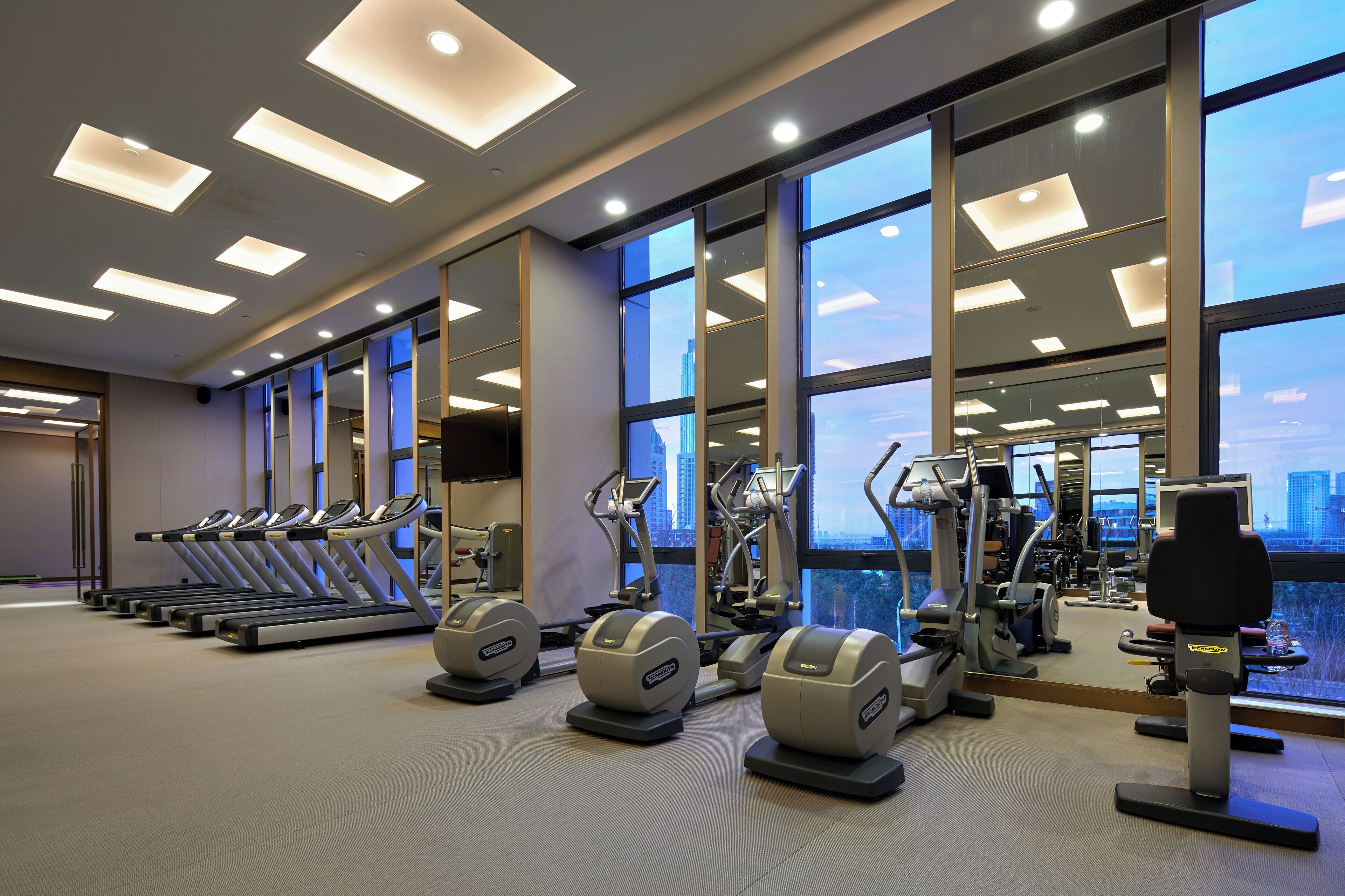 A gym with treadmills, elliptical machines, and weight equipment, large windows, and a city view.