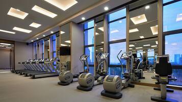 Un gimnasio con cintas de correr, elípticas y equipo de pesas, ventanas amplias y vistas a la ciudad.
