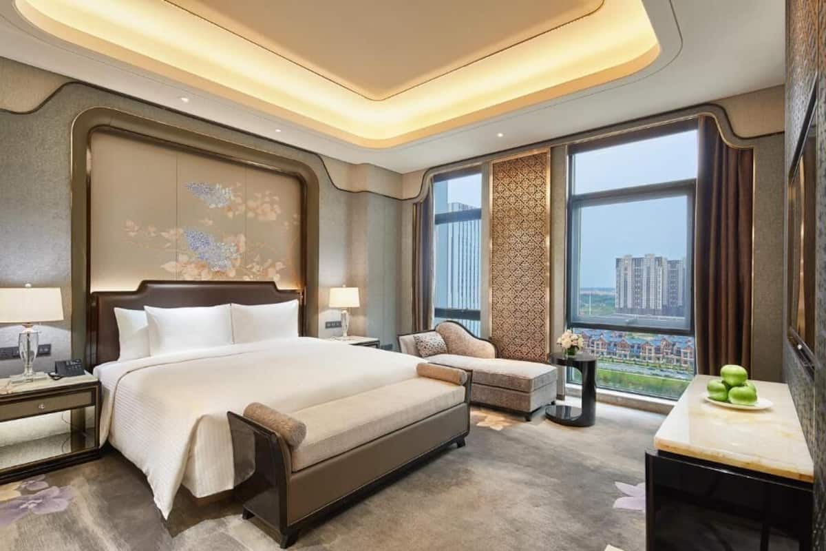 A modern hotel room with a large bed, a seating area, a small table, and a view of the city through large windows.