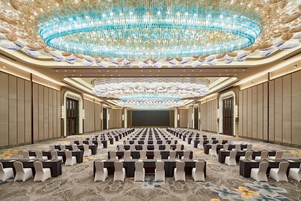 A large conference room with rows of tables and chairs, a grand chandelier, and a stage at the front.