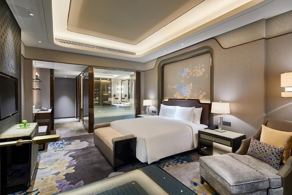 Una habitación de hotel moderna con una cama grande, una zona de estar con sofá y sillón, un escritorio con lámpara y un espejo amplio.