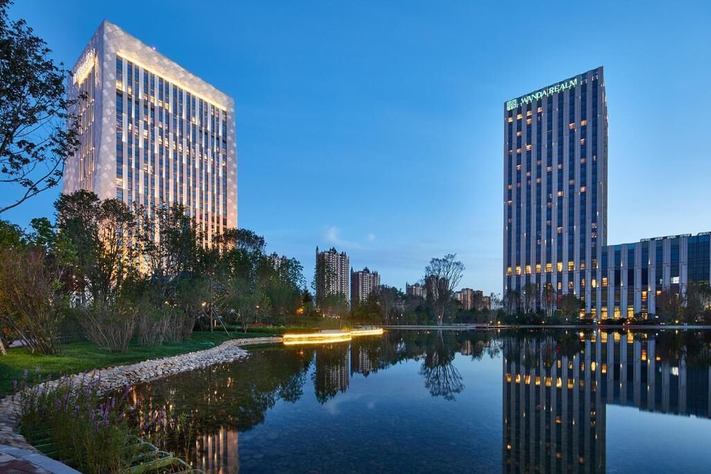 A modern building with "WandaRealm" signage, surrounded by a calm water body reflecting the structure, with other high-rise buildings in the background.