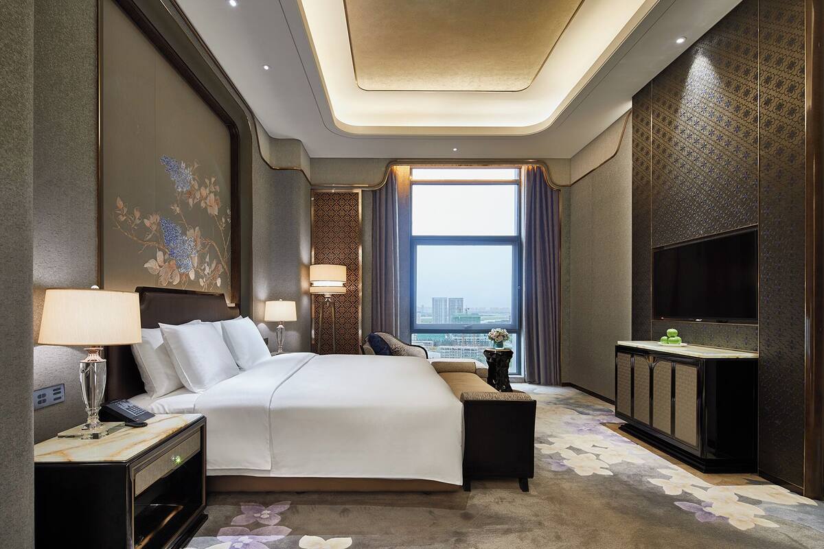 A modern hotel room with a large bed, bedside tables, a desk, a flat-screen TV, and a view of the city.