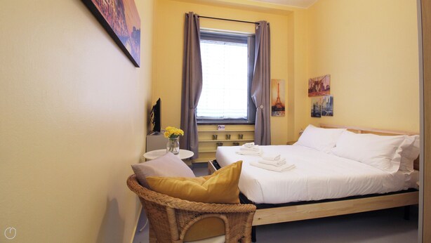Een hotelkamer met een bed, een kleine tafel met een bloevas, een rieten stoel en kunst aan de muren.