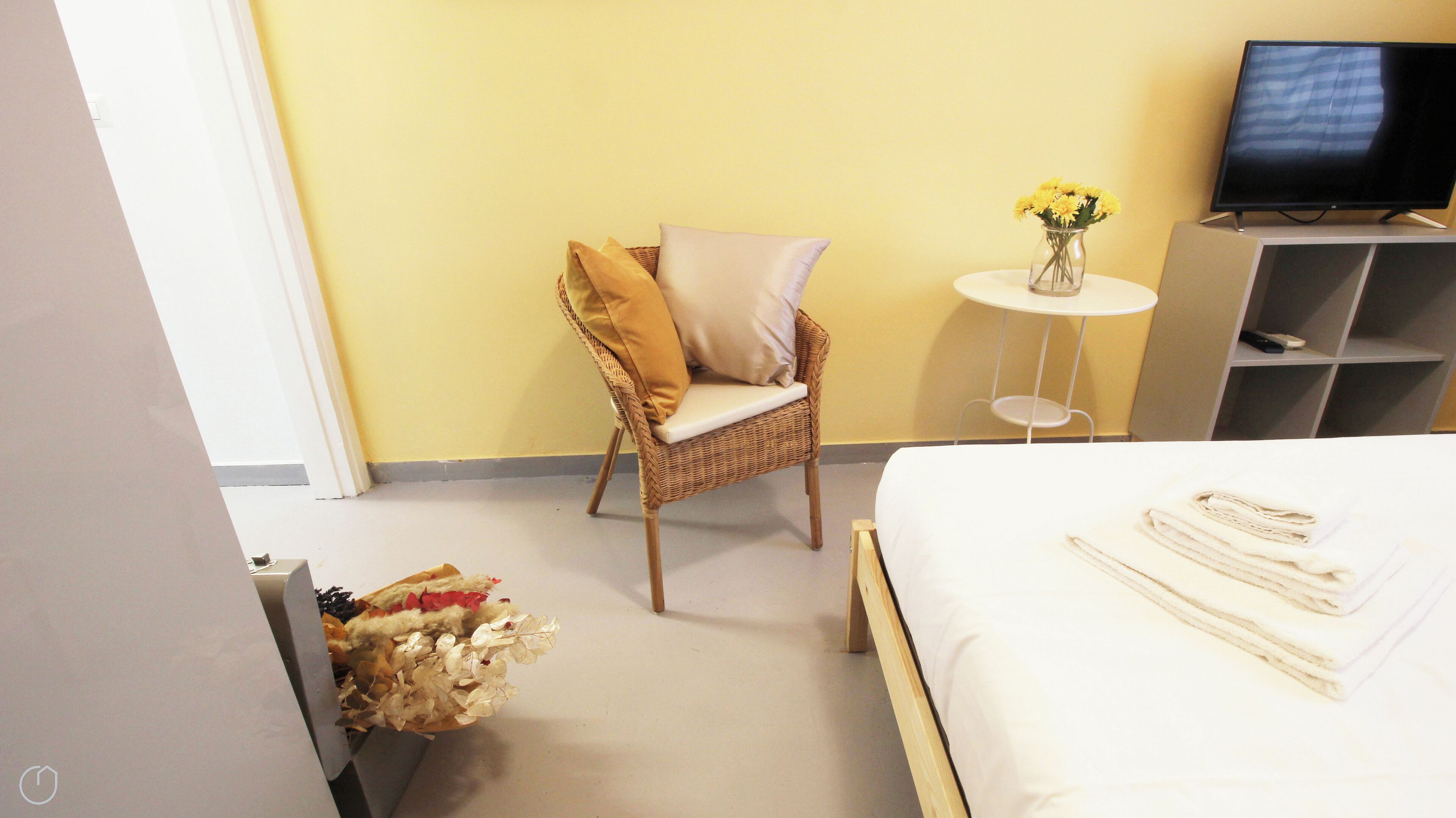 Una habitación de hotel con una cama, una silla de mimbre con cojines, una mesita redonda con un florero y un televisor sobre un mueble.