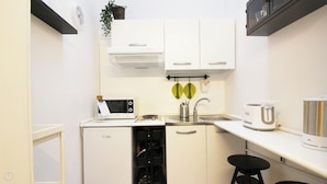 Een compacte keuken met witte kastjes, een magnetron, een spoelbak met kraan, een waterkoker en een klein krukje.