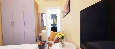 A hotel room with a bed, a TV, a wardrobe, a chair, a small table with a vase of flowers, and a window with curtains.