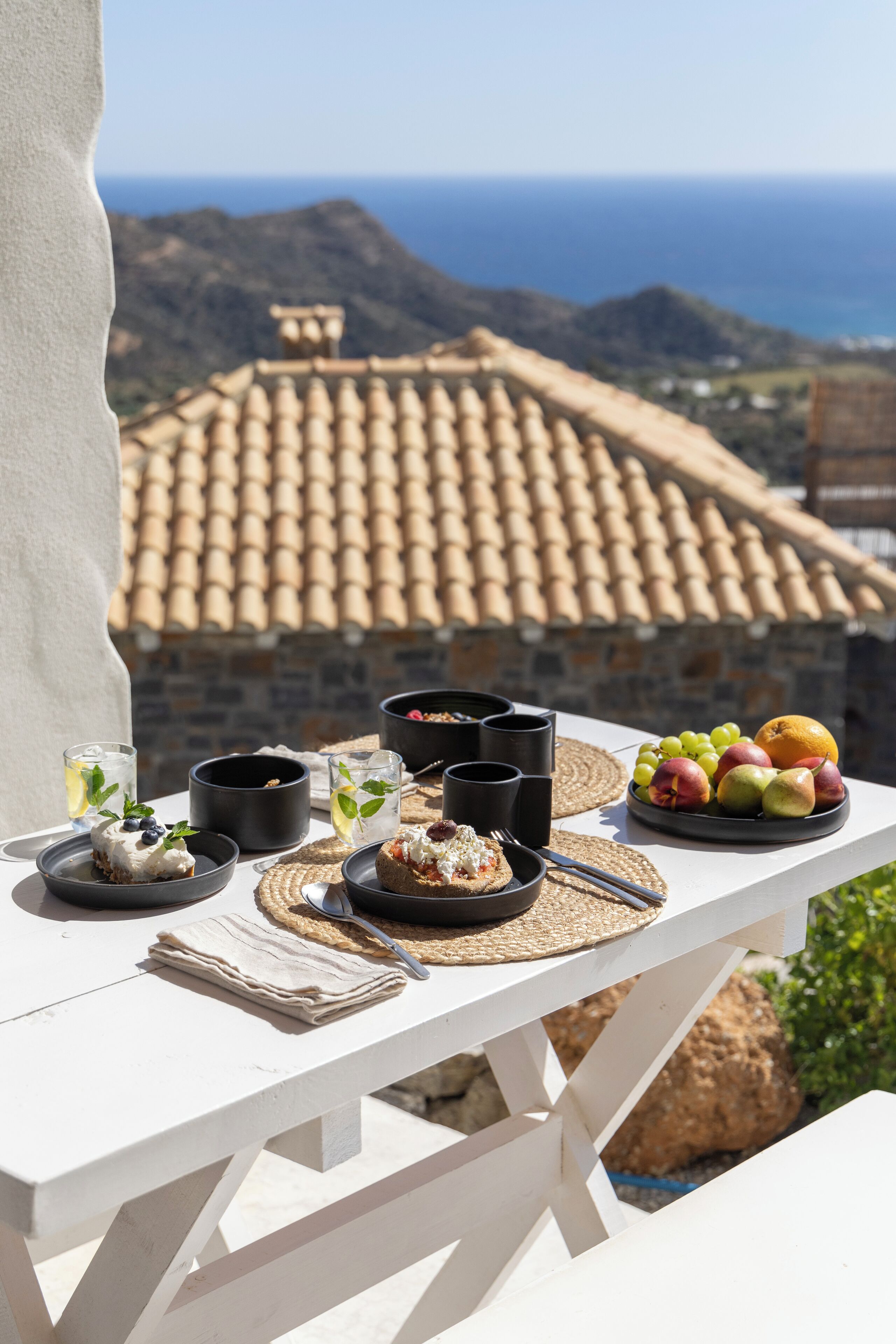 A white wooden table set with a fruit plate, black cups, and a dessert with mint garnish, overlooking a tiled-roofed building and a mountainous landscape.