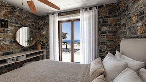 Un dormitorio con pared de piedra, una cama grande con ropa de cama blanca, un espejo redondo y vista al mar a través de un ventanal.