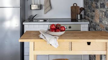 A kitchen with white cabinets, a stainless steel refrigerator, a wooden table with a bowl of fruit, and a stone wall.