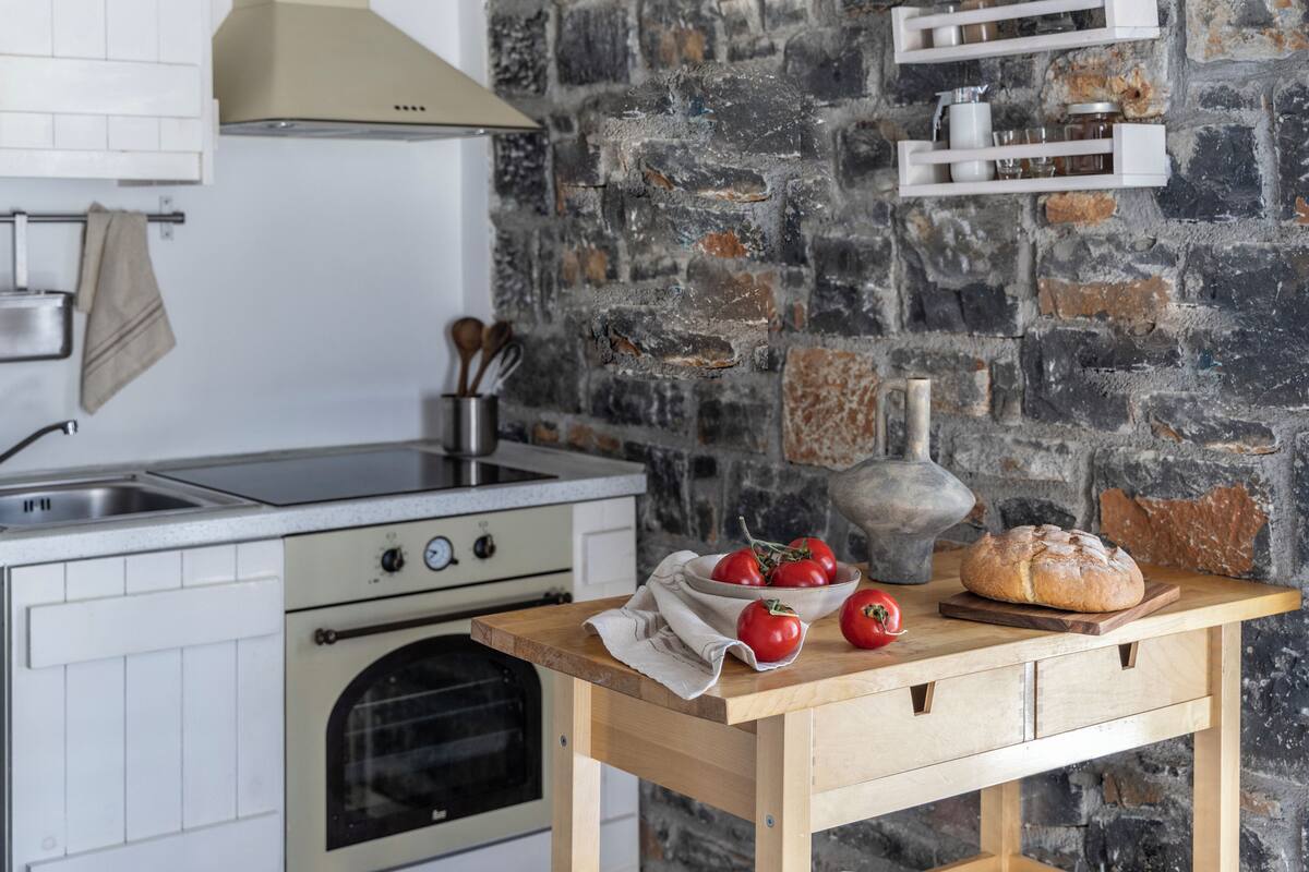 Una cocina con pared de piedra, una mesa de madera con tomates y pan, y un colgador de ollas.
