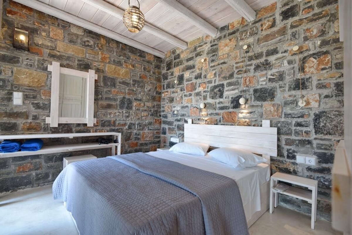 Un dormitorio con pared de piedra, cabecera de madera, cama con ropa de cama blanca, mesita de noche y lámpara colgante.