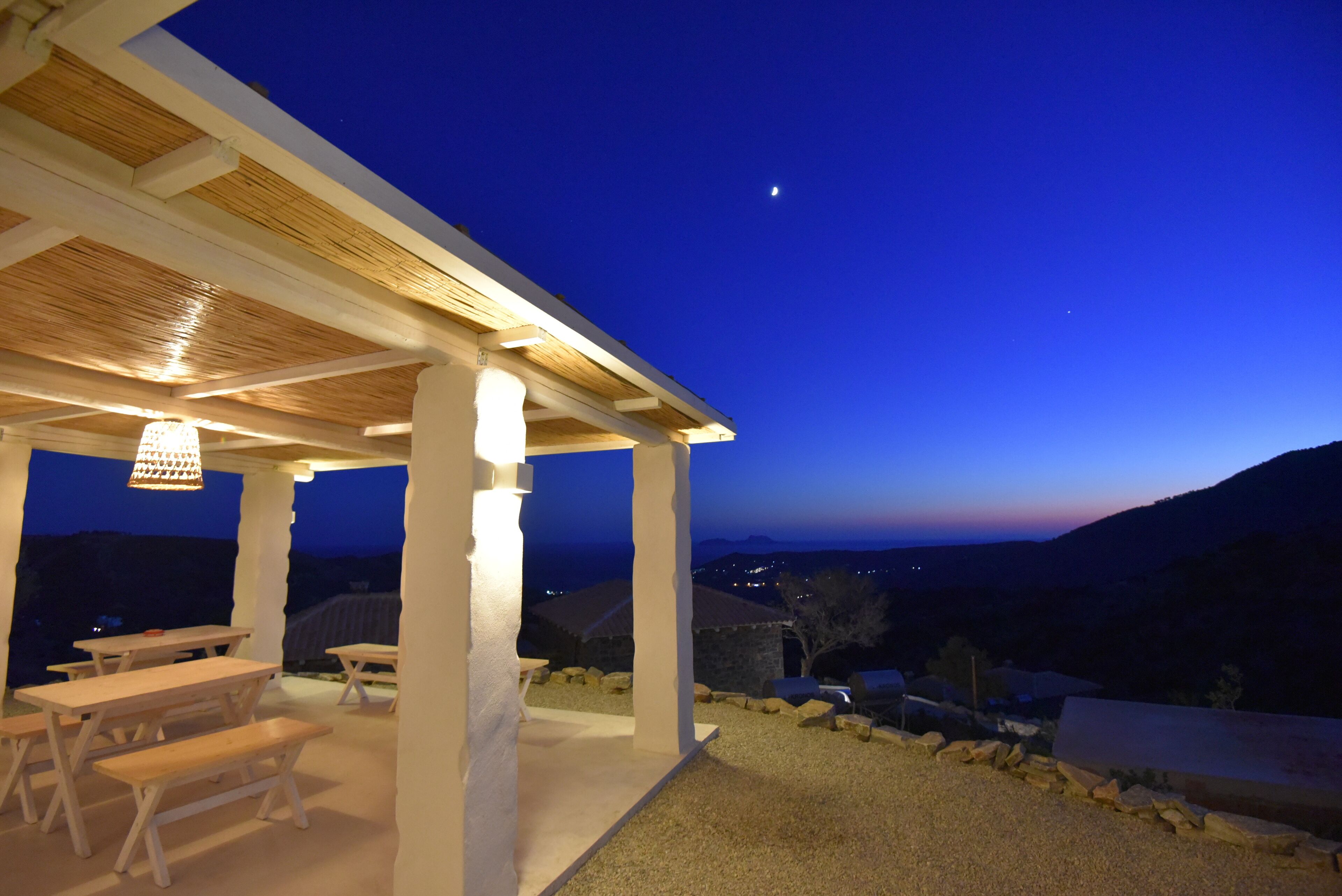 Un patio al aire libre con bancos de madera y una pérgola, con vistas a un paisaje montañoso al anochecer.