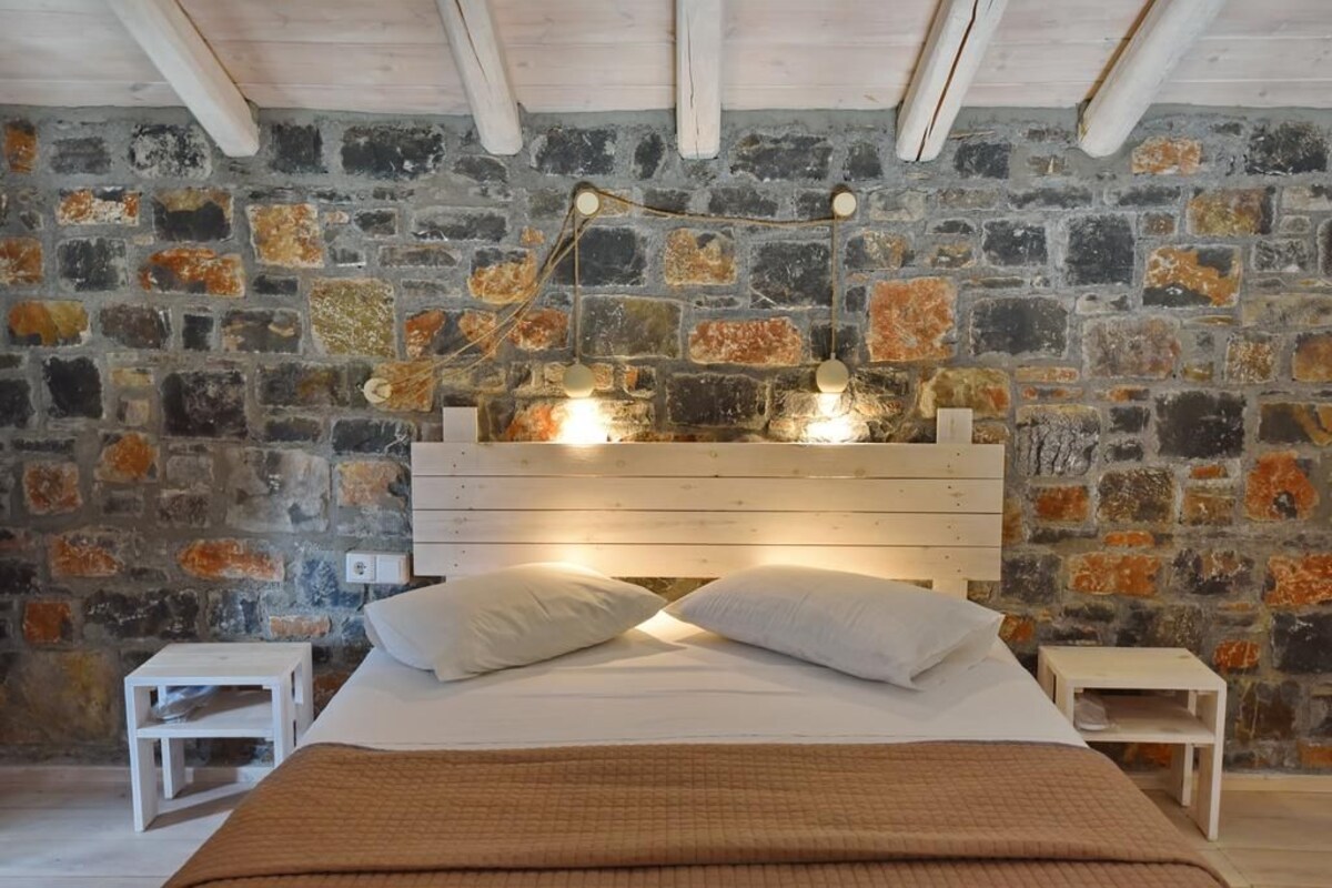 Un dormitorio con pared de piedra, cabecero de madera, dos mesitas de noche y una cama con almohadas.