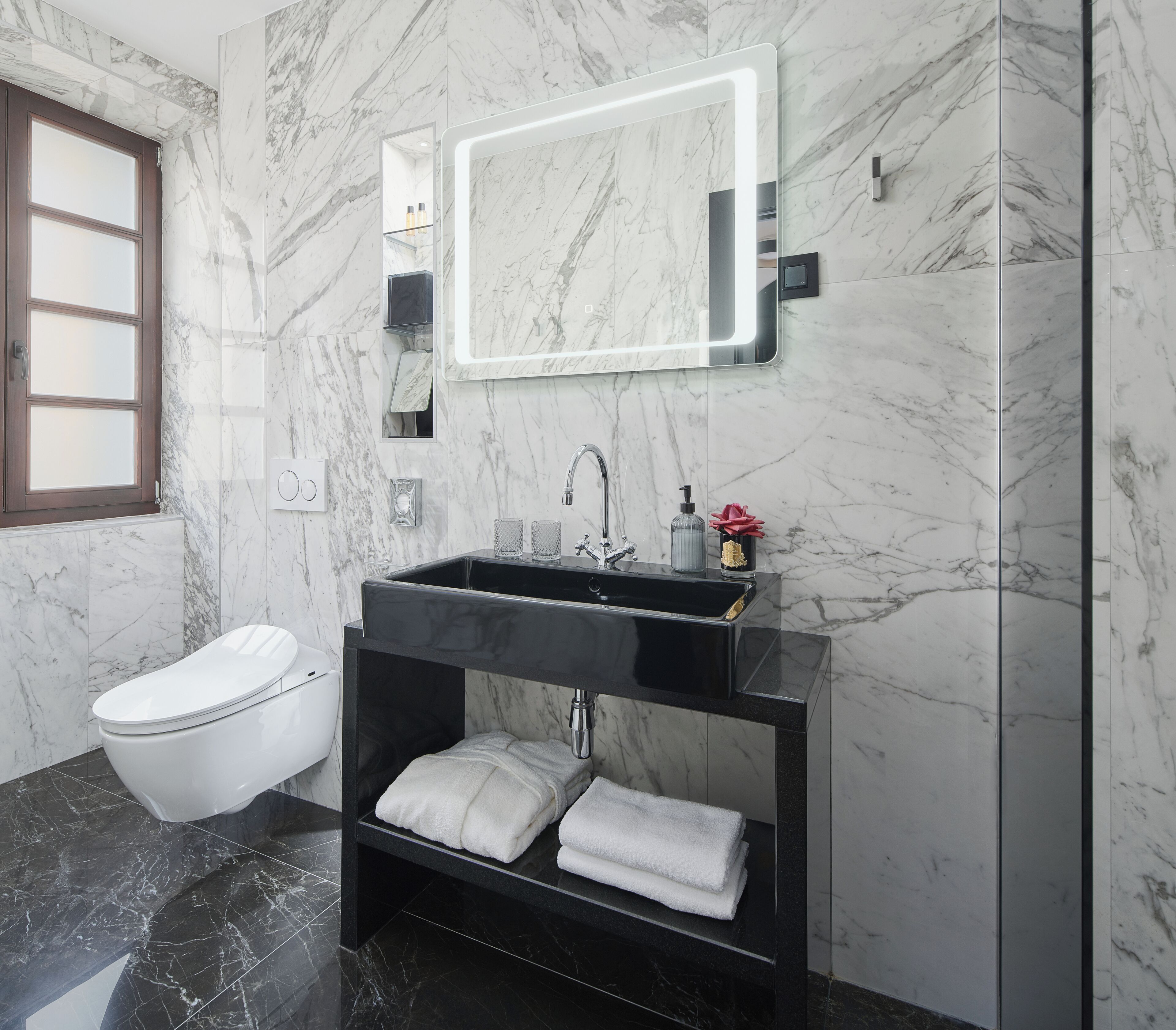 A modern bathroom with a black sink, white marble walls, a toilet, and a mirror with integrated lighting.