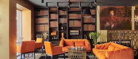 Ein modernes Wohnzimmer mit einem großen orangefarbenen Sofa, passenden Sessel und einem Couchtisch. Ein Bücherregal voller Bücher und ein Gemälde an der Wand sind ebenfalls zu sehen.